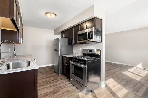 Modern kitchen with stainless steel appliances, dark cabinets, a gas stove, built-in microwave, and light wood flooring, adjacent to an open living area.