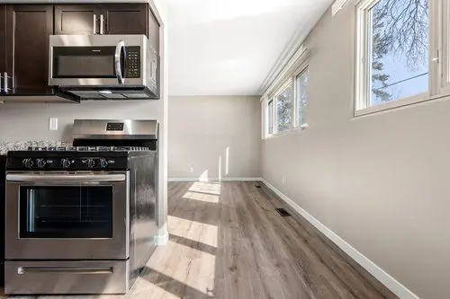Modern kitchen with stainless steel stove and microwave, dark cabinets, and an adjacent empty room with light wood flooring and large windows.