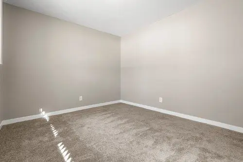 Empty room with beige carpet, light beige walls, white baseboards, and two electrical outlets. Sunlight enters through a window, casting shadows on the floor.