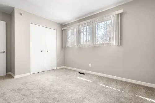 Empty, carpeted room with beige walls, closed white closet doors, and two windows with vertical blinds letting in natural light.