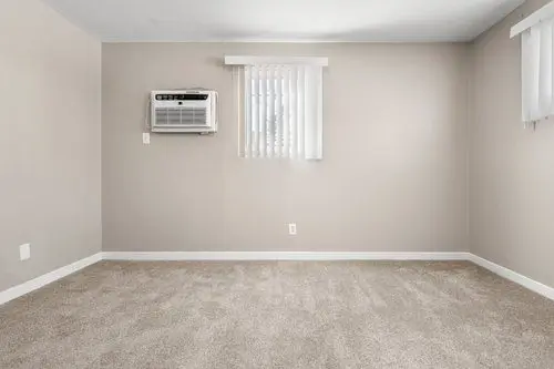 Empty room with beige carpet, light-colored walls, a window with vertical blinds, and a wall-mounted air conditioning unit.