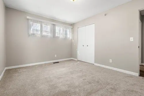 Empty room with beige carpet, light-colored walls, a set of three windows with sheer curtains, and a closed white double-door closet.