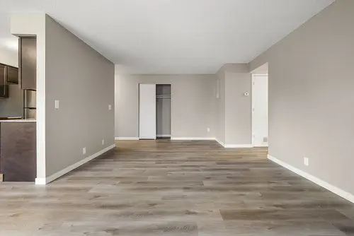 Empty room with light gray walls, wood flooring, an open closet, and a partial view of a kitchen on the left.
