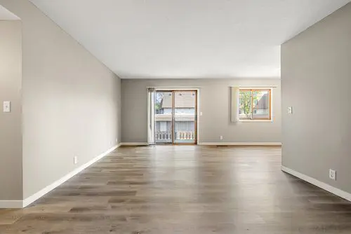 Empty living room with light-colored walls, wood flooring, a sliding glass door leading to a balcony, and a window letting in natural light.