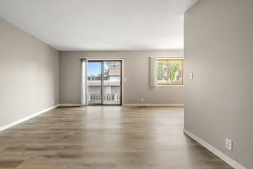 Empty living room with light wood flooring, beige walls, a sliding glass door leading to a balcony, and a window letting in natural light.