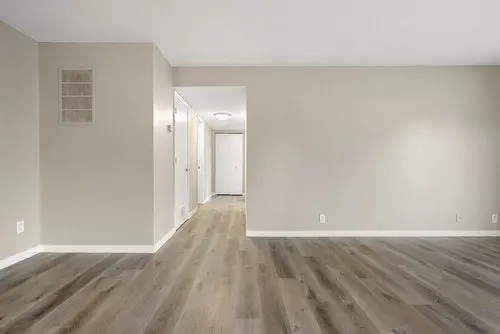 Empty room with light beige walls, wood laminate flooring, and a hallway leading to a closed door in the background.