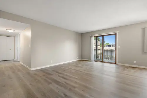 Empty living room with light wood floors, beige walls, and a sliding glass door leading to a balcony with a view of neighboring buildings.