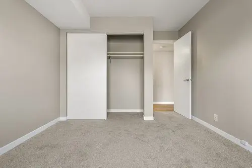 Empty beige bedroom with carpet flooring, an open closet with sliding doors, and an open door leading to another room.