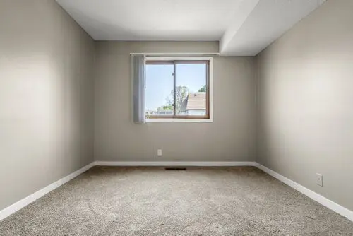 Empty room with beige walls, carpeted floor, and a single window with partial blinds letting in natural light.