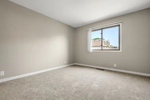 Empty room with beige carpet, light gray walls, and a window with a view of neighboring buildings, letting in natural light.