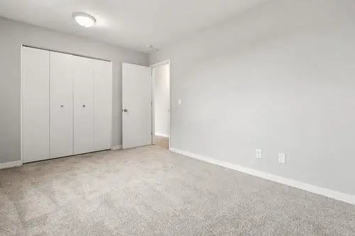 Empty room with light gray walls, beige carpet, a closed white closet, an open door, ceiling light, and electrical outlets on one wall.