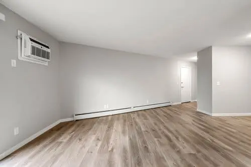 Empty room with light gray walls, wood-style flooring, baseboard heating, and a window air conditioning unit mounted on the wall.