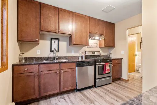 Modern kitchen with wood cabinets, stainless steel appliances, granite countertops, and a wood-look floor. A bathroom is visible through an open door in the background.