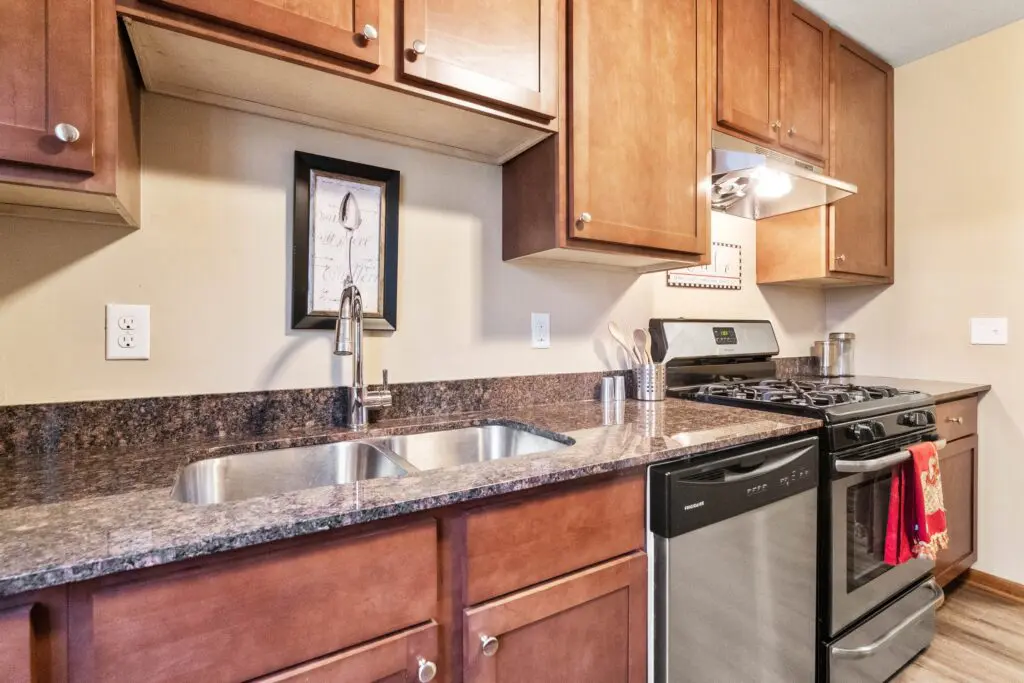 Modern kitchen with brown cabinets, granite countertops, double sink, stainless steel dishwasher, and a gas stove with a red towel hanging on the oven door.