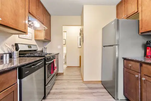 A modern kitchen with stainless steel appliances, wooden cabinets, and light wood flooring. A refrigerator and dishwasher are visible, along with various kitchen utensils on the counter.