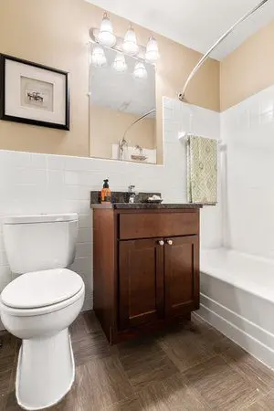 A small bathroom with a toilet, wooden vanity with sink, wall mirror, framed picture, towel rack, and a shower-tub combo with a curved shower rod.