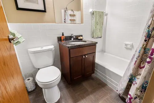 A small bathroom with a toilet, dark wood vanity with sink, shower-tub combo, white tiles, and patterned shower curtain.
