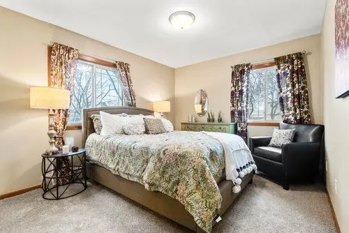 A bedroom with a double bed, floral bedding, two windows with patterned curtains, a black armchair, lamps, and a green dresser with a mirror.