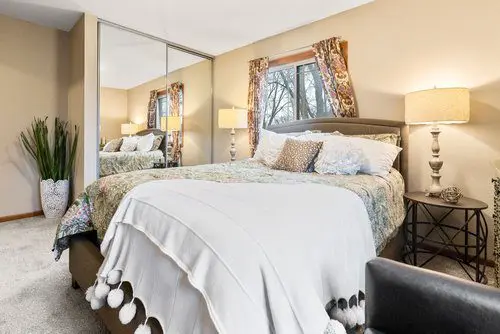 A neatly made bed with patterned bedding and decorative pillows in a bedroom with beige walls, mirrored closet doors, and two lamps on bedside tables.