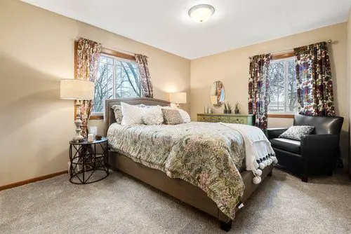 A bedroom with a double bed, floral bedding, two windows with patterned curtains, a nightstand, a green dresser with a mirror, and a black armchair.