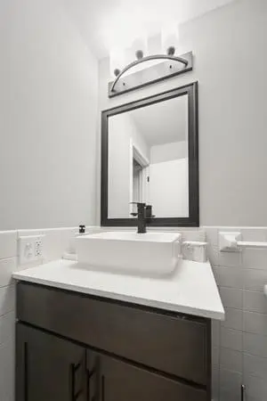 A modern bathroom vanity with a rectangular sink, black faucet, large framed mirror, and wall-mounted light fixture above. White tiled backsplash and countertop are visible.