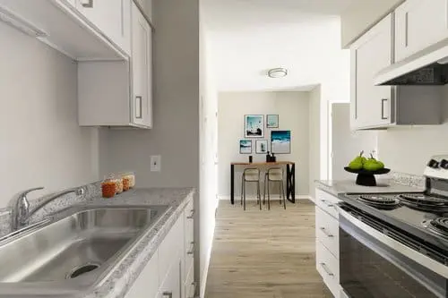 Modern kitchen with white cabinets, stainless steel sink, electric stove, and a view of a small dining area with two chairs and framed wall art in the background.