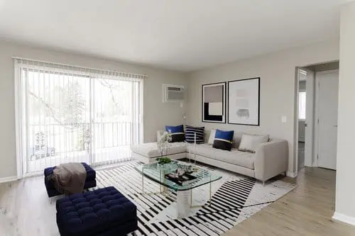 A modern living room with a beige sectional sofa, navy ottomans, a glass coffee table, striped rug, and large windows with vertical blinds.