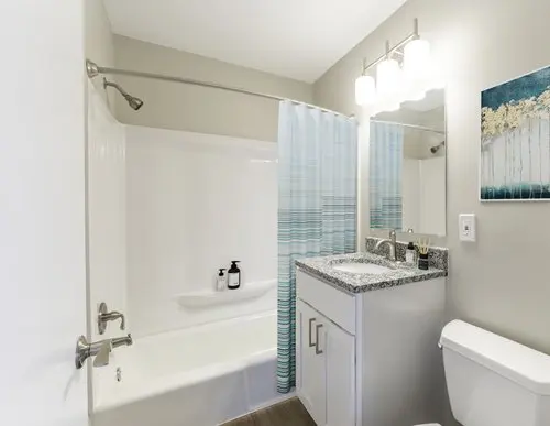 A modern bathroom with a white bathtub, blue striped shower curtain, granite vanity, mirror, and toilet. Two soap dispensers are on the tub ledge, and a painting hangs on the wall.
