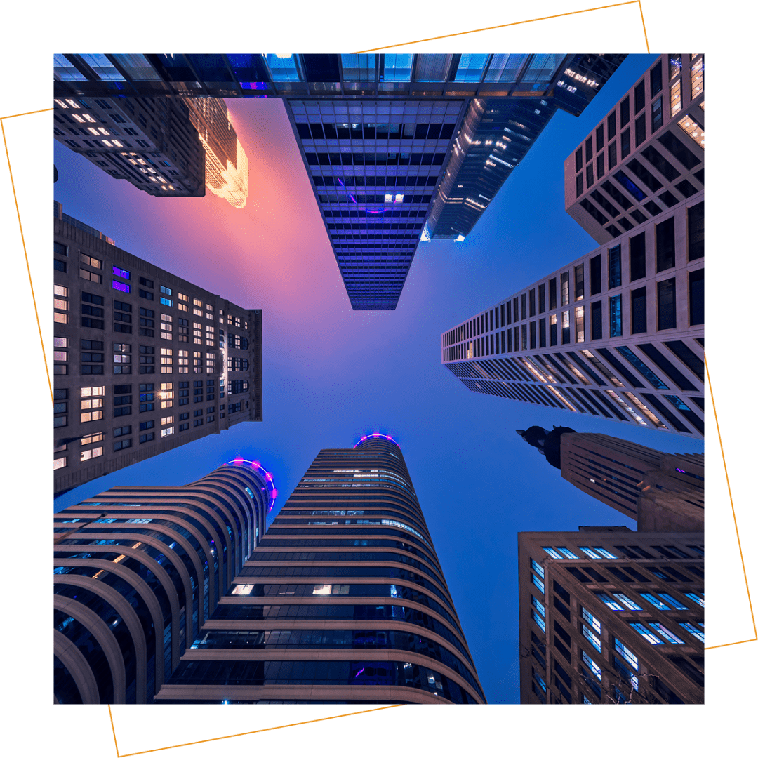 View of tall modern skyscrapers taken from the ground looking up at dusk, with building lights on and a gradient sky in the background.