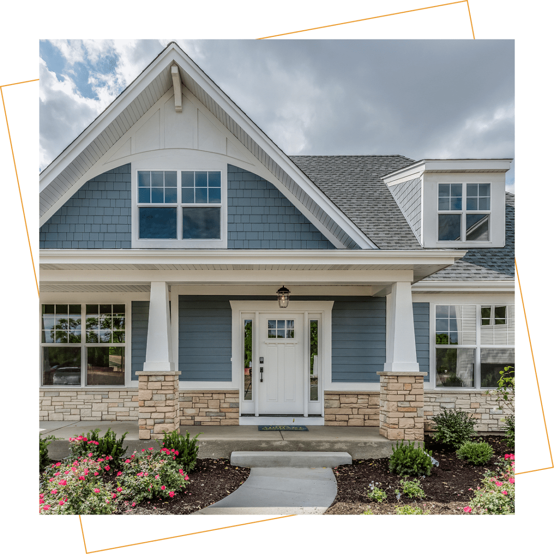 A modern suburban house with blue siding, stone accents, white trim, and a covered front porch, surrounded by landscaped bushes and flowers.