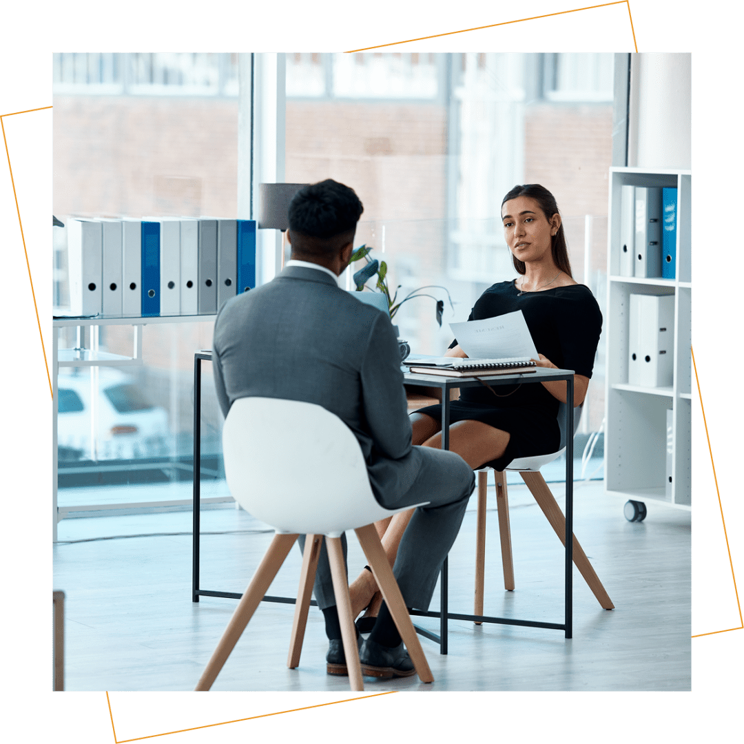 Two people sit across from each other at a small table in a modern office, engaged in a professional conversation with documents in hand.
