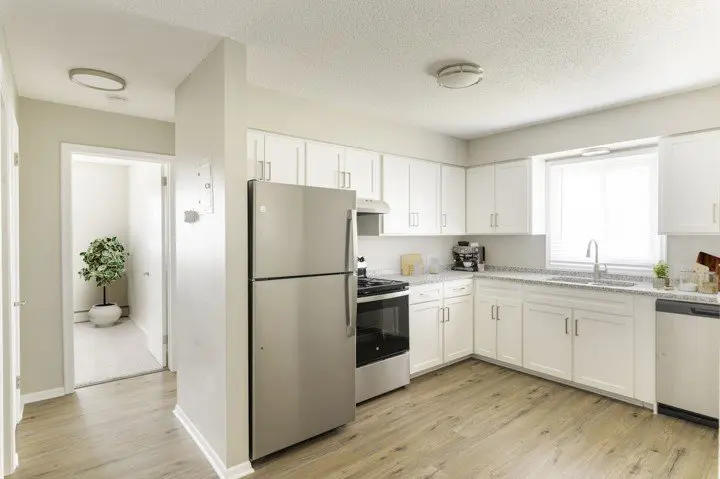 Modern kitchen with white cabinets, stainless steel appliances, wood-style flooring, and a window above the sink; hallway with a potted plant visible in the background.