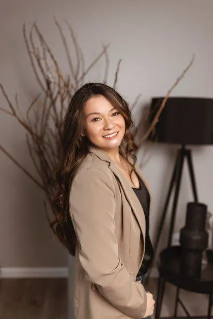 Young woman with long brown hair wearing a beige blazer and black top, standing indoors in front of decorative branches and a black lamp, smiling at the camera.