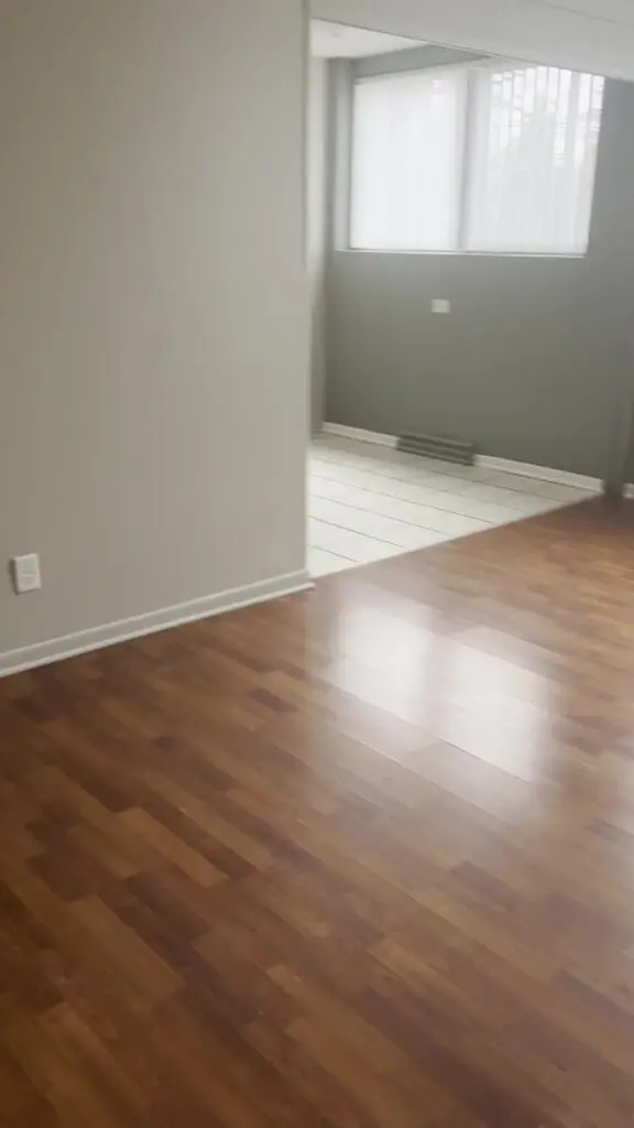 Empty room with wood flooring leading to a tiled area with large windows and vertical blinds. Pale gray walls and a wall outlet are visible.