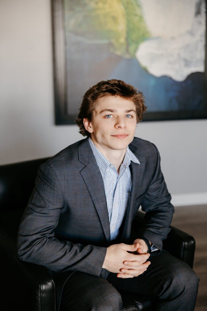 A young man in a gray suit sits on a black chair, hands clasped, looking toward the camera with a neutral expression. There is abstract art on the wall behind him.