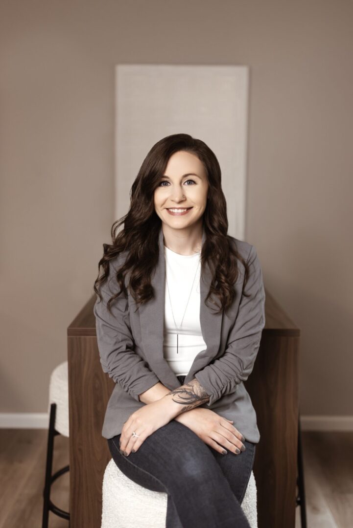 A woman with long brown hair sits on a stool, wearing a gray blazer, white shirt, and jeans, smiling at the camera in a neutral-toned room.