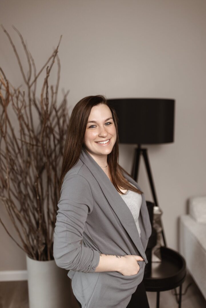 A woman with long brown hair wearing a grey cardigan smiles while standing indoors, with a black lamp and decorative branches in the background.