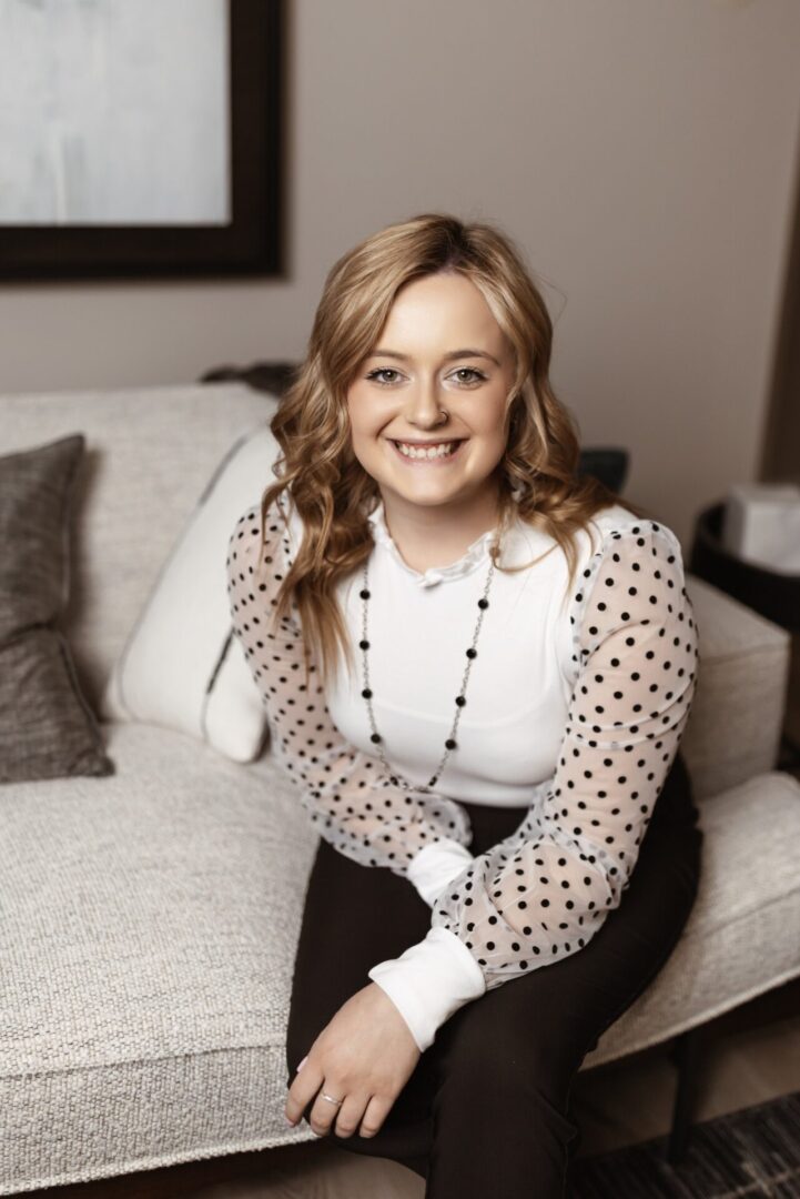A young woman with wavy blonde hair, wearing a white top with sheer polka-dot sleeves and black pants, sits smiling on a light-colored sofa.