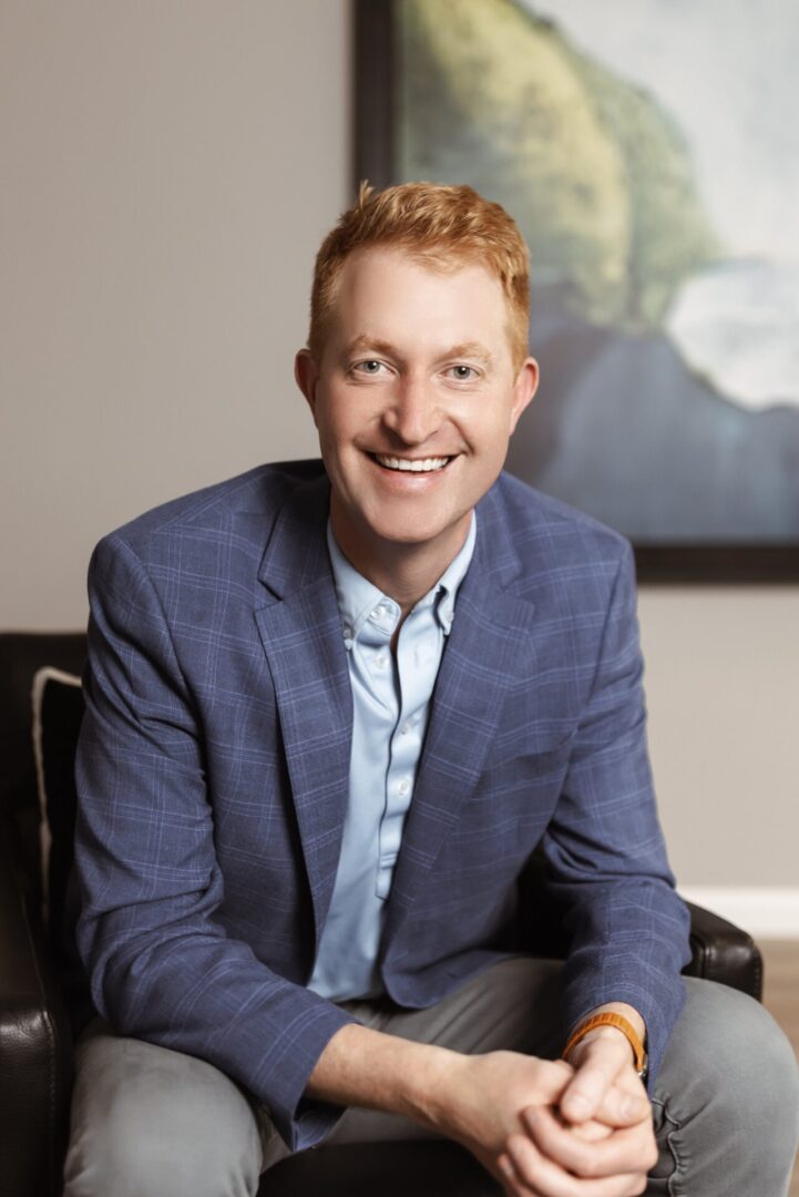 A man with short red hair, wearing a blue plaid blazer and light blue shirt, sits smiling on a black chair in front of a framed landscape painting.