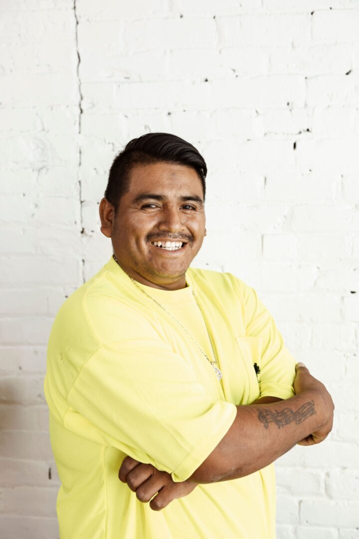 A man in a yellow shirt stands in front of a white brick wall, smiling with his arms crossed.
